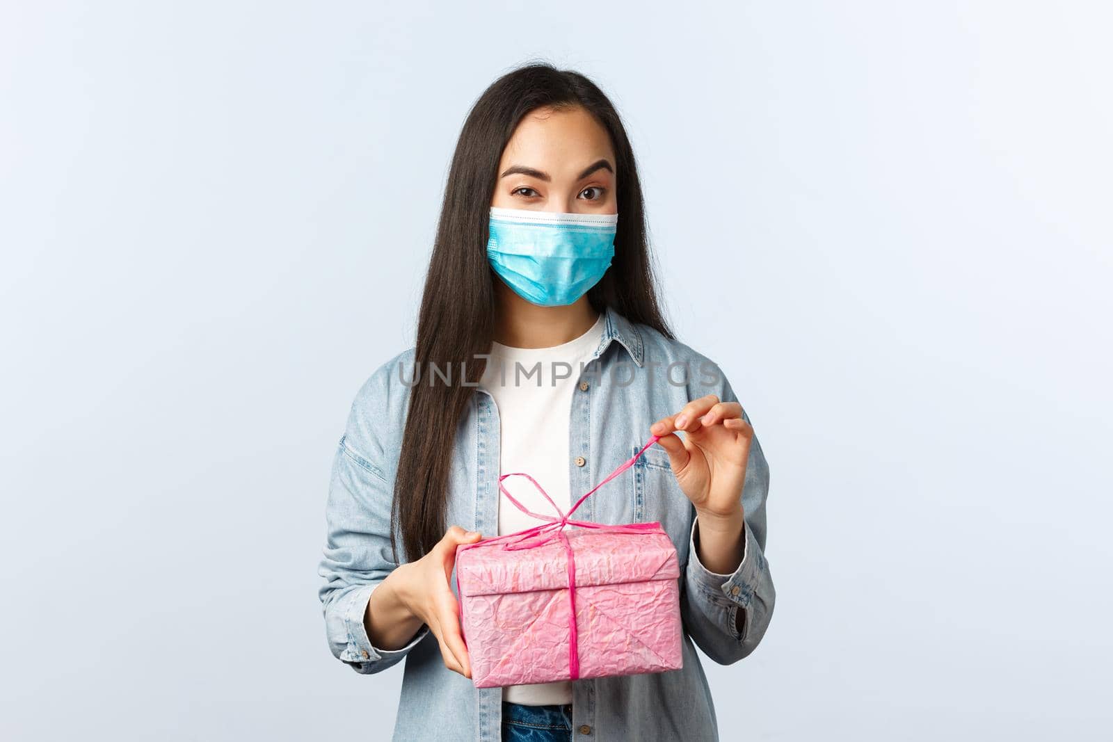 Social distancing lifestyle, covid-19 pandemic, celebrating holidays during coronavirus concept. Intrigued happy asian woman in medical mask unwrap gift box wonder what inside by Benzoix