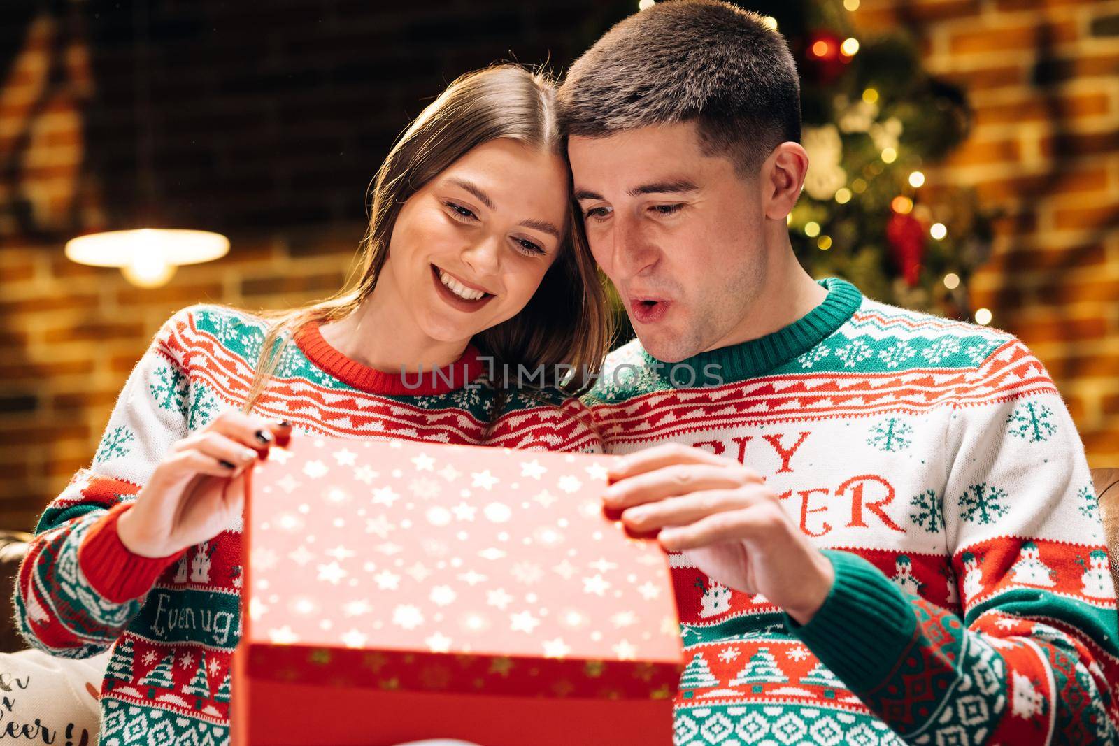 Romantic couple opening a present gift box in the evening near decorated xmas tree. Happy man is making christmas gift to his beloved woman. Concept of holidays, romance, surprise.