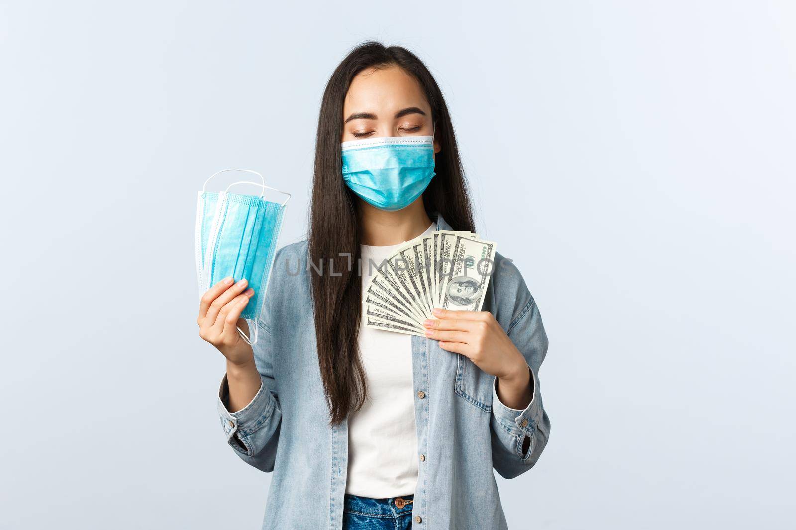 covid-19 pandemic, coronavirus expences and finance concept. Satisfied greedy asian girl in medical mask hugging money and showing masks, earn cash during coronavirus lockdown.