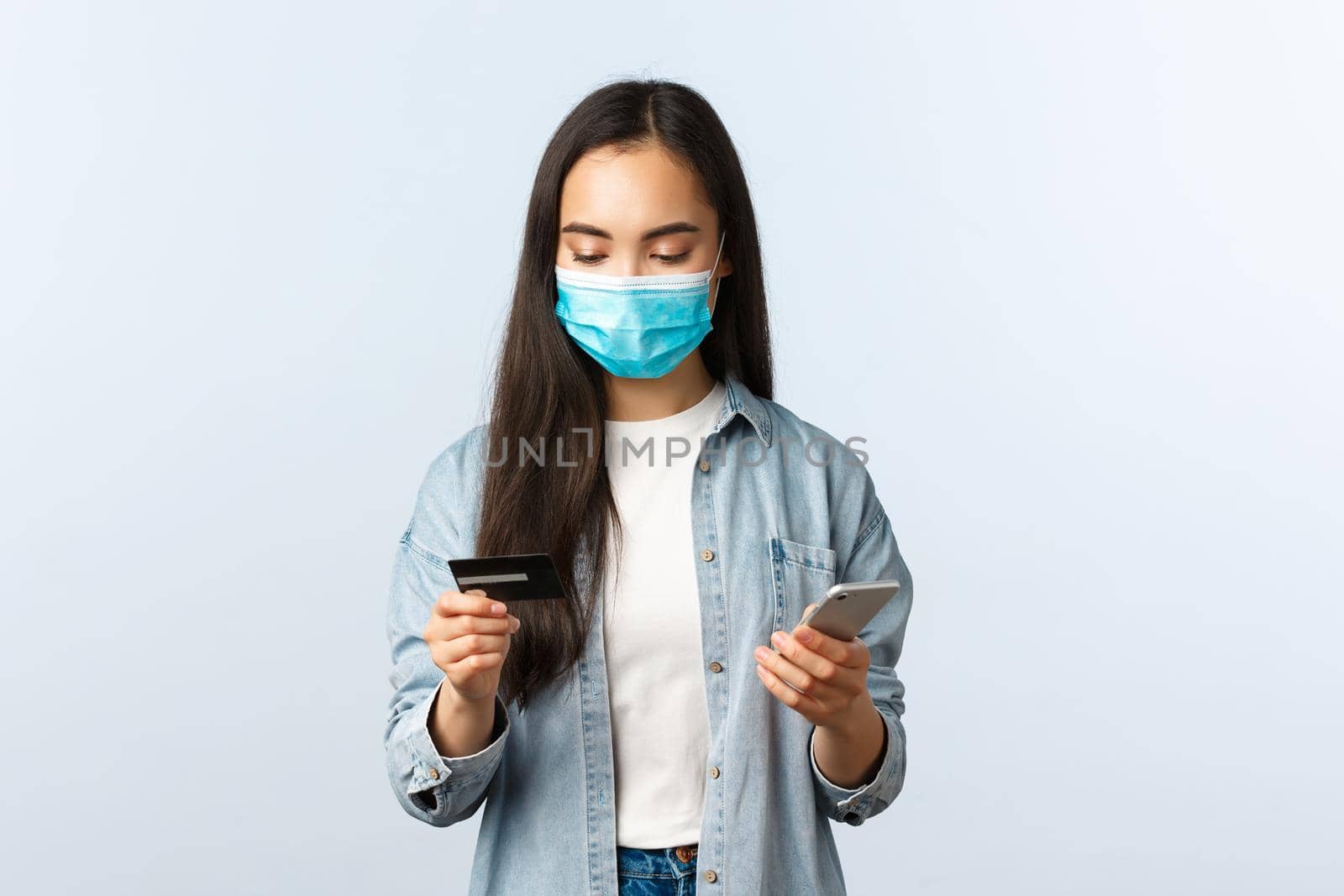 Social distancing lifestyle, covid-19 pandemic and contactless shopping concept. Attractive stylish asian girl in medical mask looking at credit card while making order on mobile application by Benzoix