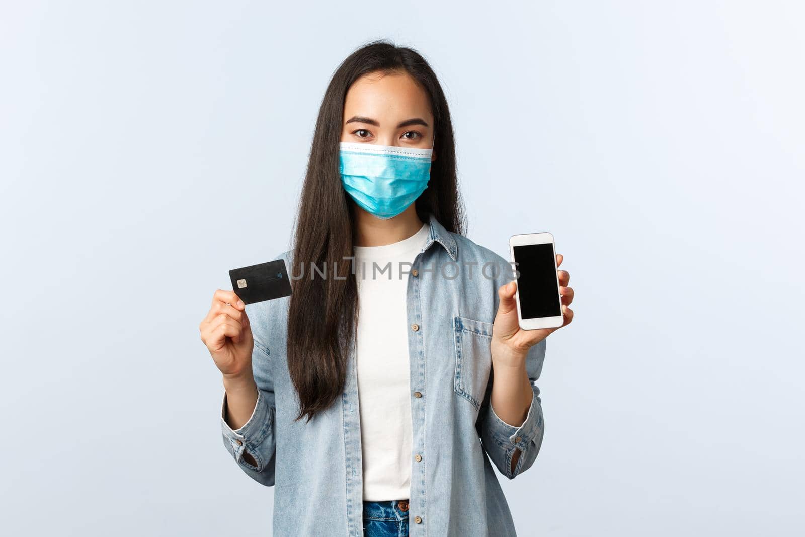 Social distancing lifestyle, covid-19 pandemic and contactless shopping concept. Young pretty asian woman in medical mask showing mobile phone screen and credit card, buying online.