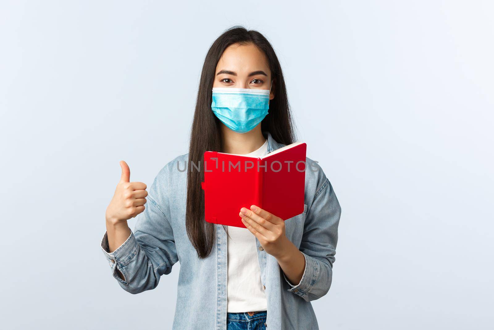 Covid-19 pandemic, education during coronavirus, back to school concept. Pleased happy asian girl in medical mask show thumb-up as checking friend homework task in notebook.