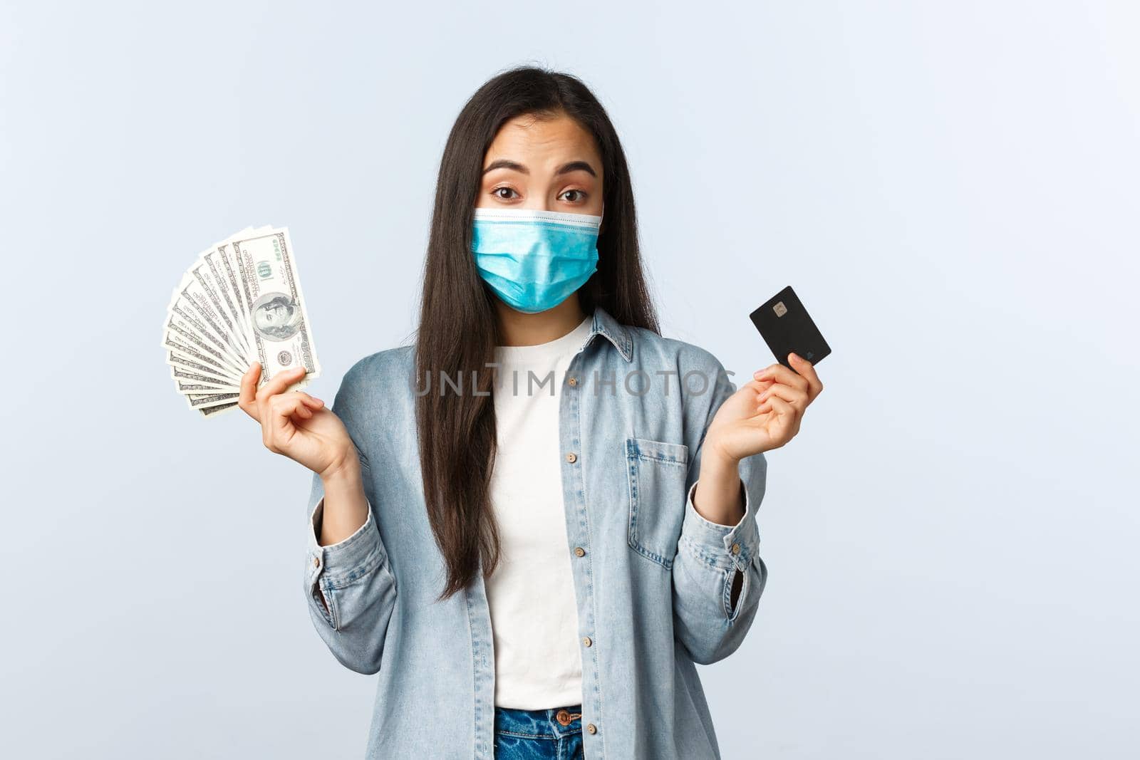 Social distancing lifestyle, covid-19 pandemic business and employement concept. Indecisive cute asian girl in medical mask shrugging shoulders as holding cash dollars and credit card.