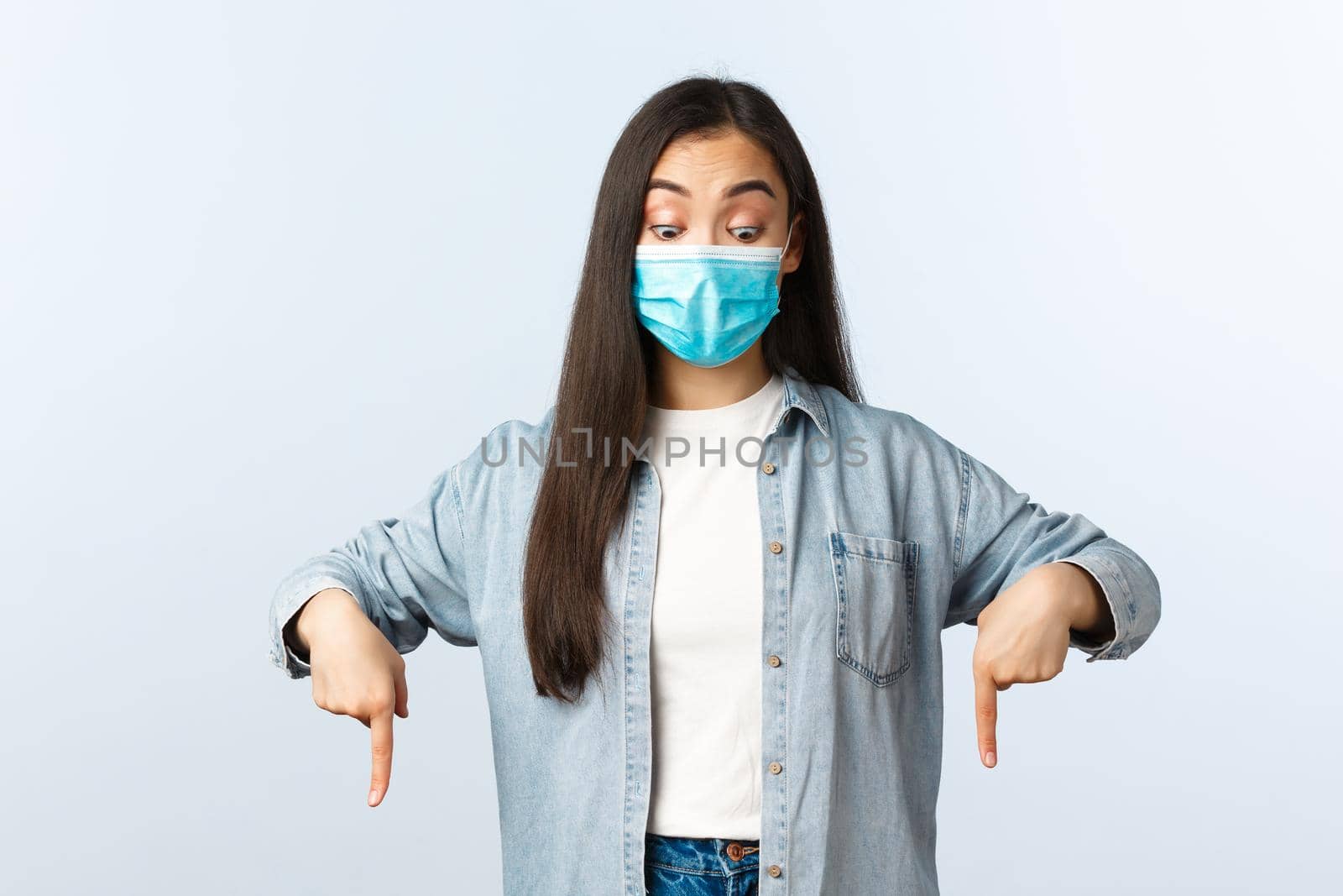 Social distancing lifestyle, covid-19 pandemic everyday life and leisure concept. Intrigued and surprised asian woman in medical mask looking, pointing fingers down at interesting banner.
