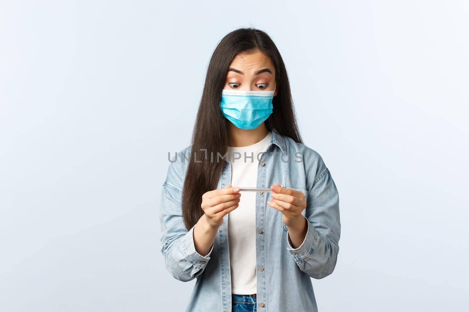Social distancing lifestyle, covid-19 pandemic everyday life and leisure concept. Shocked asian girl in medical mask measure temperature with thermometer, staring at result surprised by Benzoix