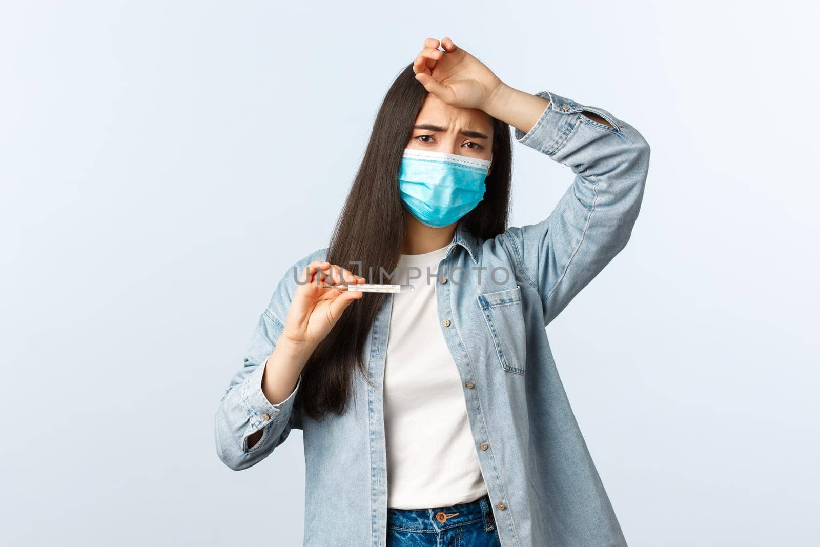 Social distancing lifestyle, covid-19 pandemic everyday life and leisure concept. Sick woman with coronavirus disease, having high fever, touch forehead and showing thermometer, wear medical mask by Benzoix
