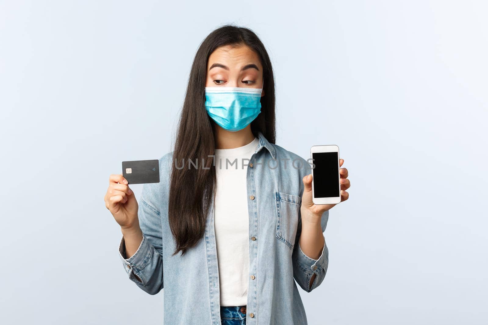 Social distancing lifestyle, covid-19 pandemic and contactless shopping concept. Intrigued cute asian girl in medical mask peeking at credit card, showing smartphone display by Benzoix