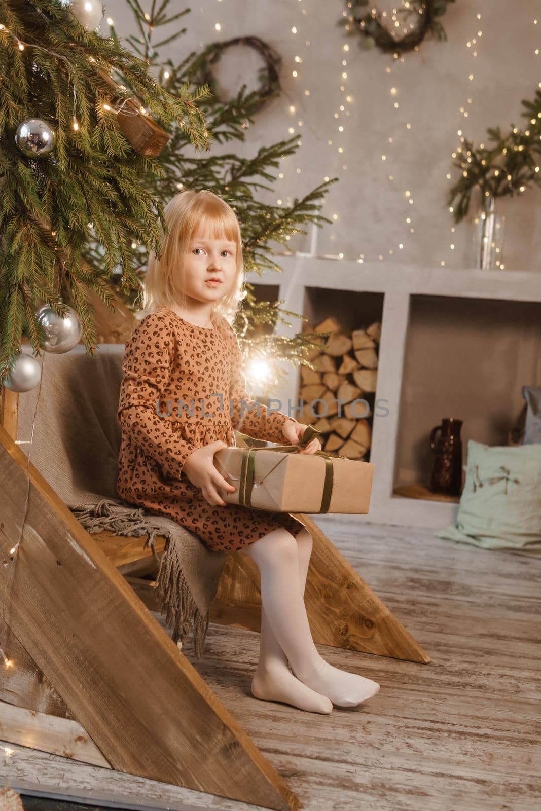 A little blonde girl is sitting on a wooden staircase in a Scandinavian interior decorated in a New Year's style. A child holds a Christmas gift in a craft package.
