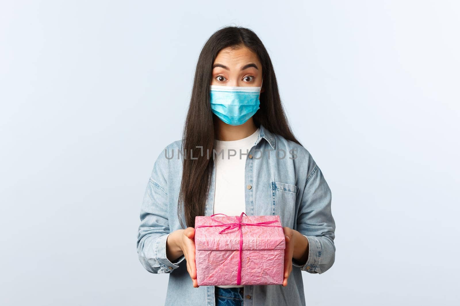 Social distancing lifestyle, covid-19 pandemic, celebrating holidays during coronavirus concept. Surprised birthday girl in medical mask receive present, looking at gift box amazed by Benzoix