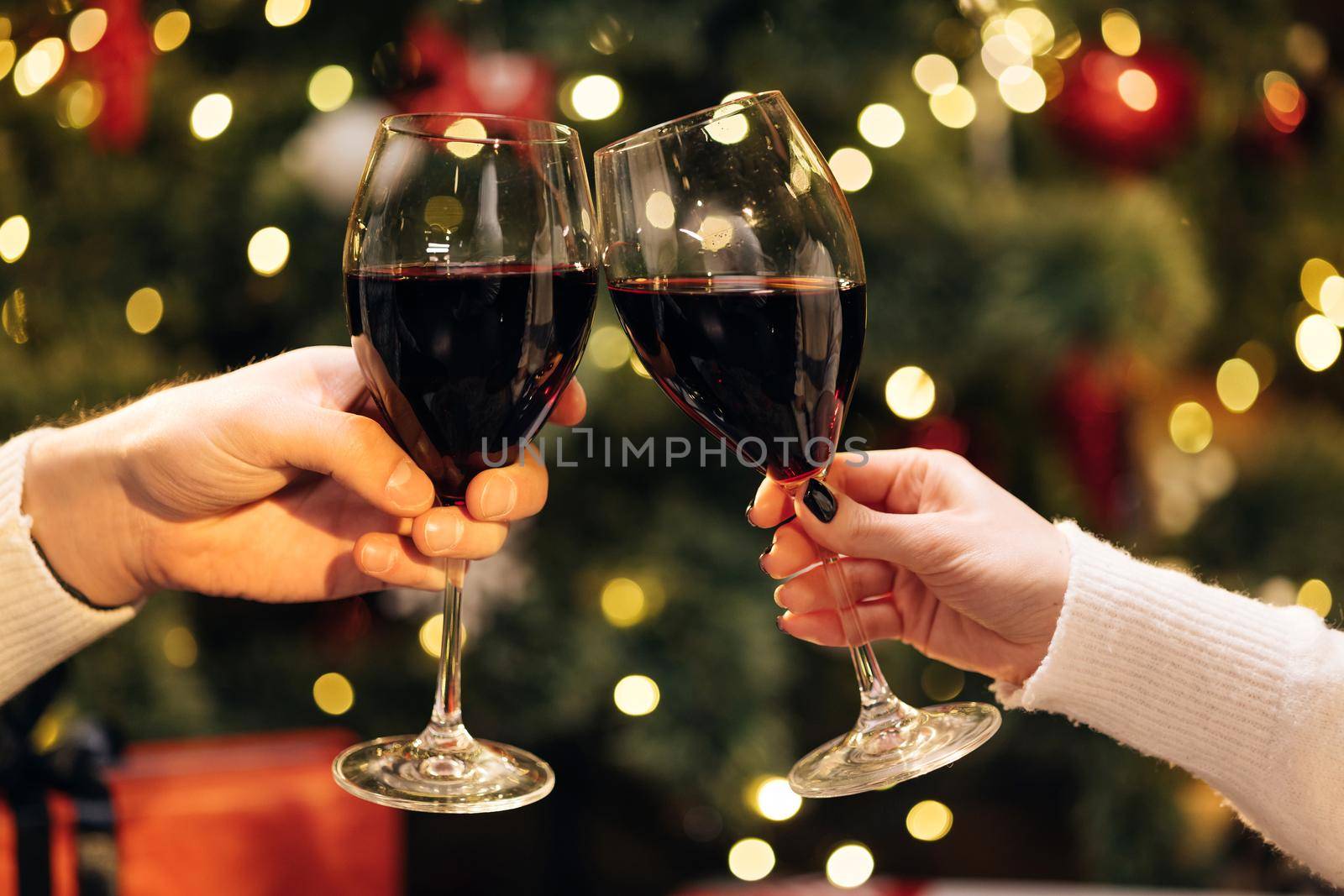Close up Hands of unknown people who cheering with glasses of wine against Christmas tree decorated with glowing garland. Blurred background, bokeh. Happy New Year.