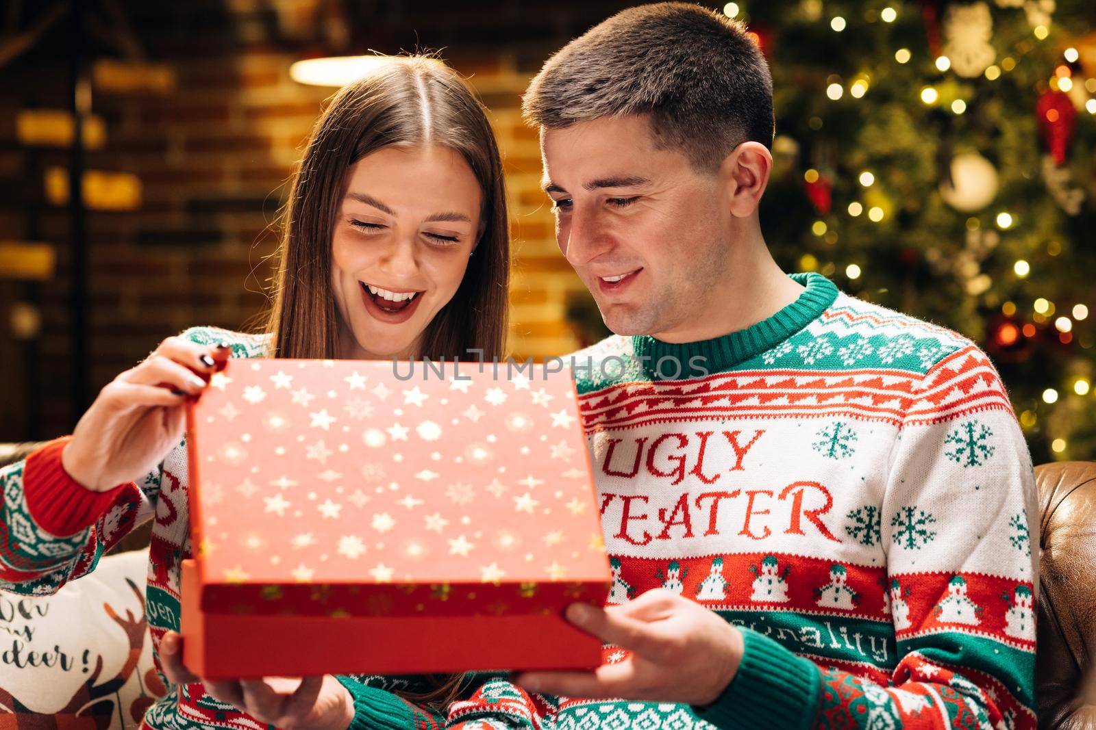 Portrait of cheerful young woman opening xmas gift box with excited surprised face feeling happy on New Year's Eve sitting at home near glowing christmas tree. Xmas concept. Holiday miracle.