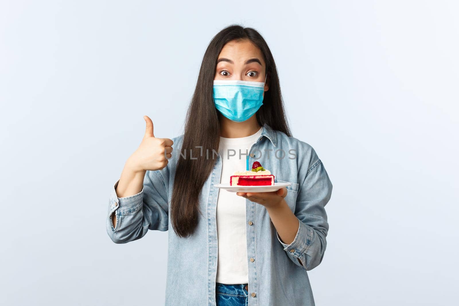 Social distancing lifestyle, covid-19 pandemic, celebrating holidays during coronavirus concept. Happy smiling asian birthday girl in medical mask, show bday cake and thumb-up pleased.