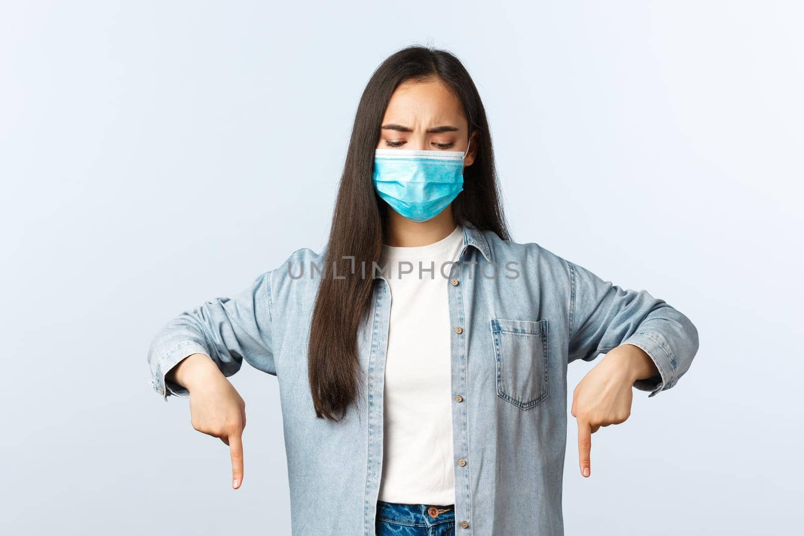 Social distancing lifestyle, covid-19 pandemic everyday life and leisure concept. Serious concerned asian woman in medical mask, looking and pointing down with displeased face.