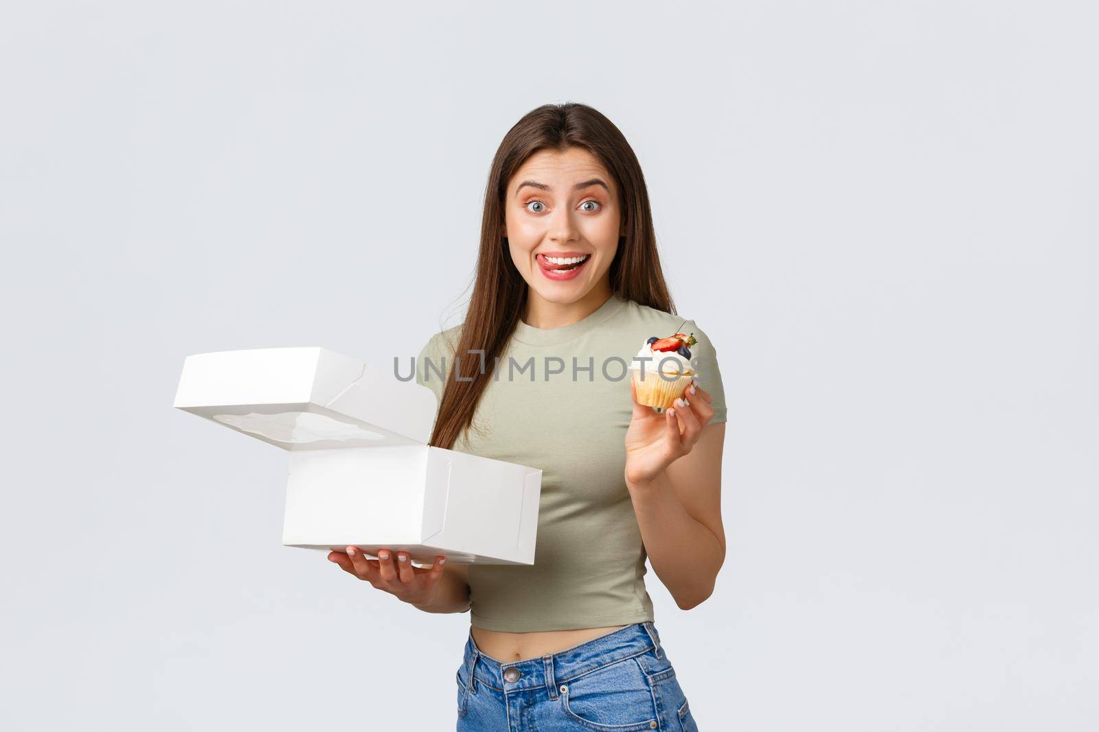 Delivery, lifestyle and food concept. Excited smiling young woman ordered box with sweets from favourite cafe or pastry shop, holding delicous cupcake, lick lips from desire.