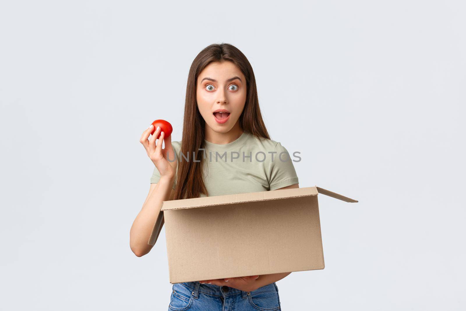 Online home delivery, internet orders and grocery shopping concept. Excited cute female customer receive groceries from internet store, take-out tomato and smiling amused.