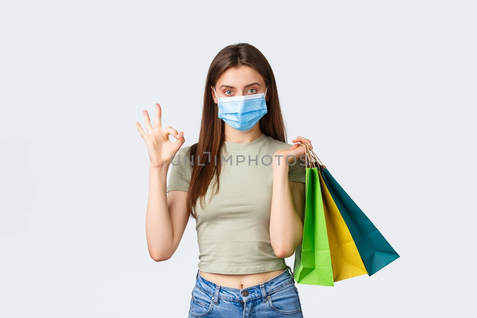 Social distancing, coronavirus and shopping, fashion concept. Stylish good-looking woman in medical mask holding shopping bags and show okay sign as recommend store in mall, white background.