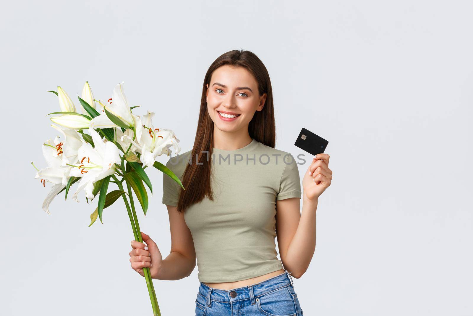 Happy tender young woman ordered bouquet of flowers delivery with credit card, smiling pleased and holding beautiful white lilies. Girl paying for purchase in internet, standing white background by Benzoix