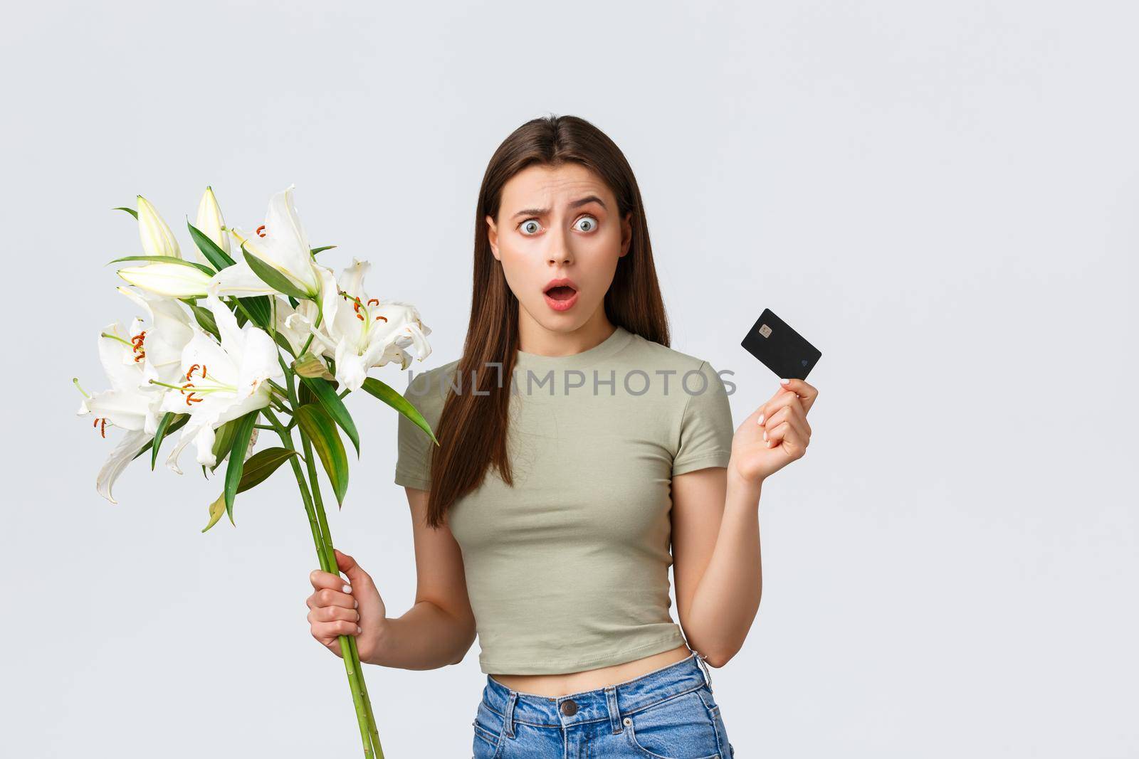 Happy tender young woman ordered bouquet of flowers delivery with credit card, smiling pleased and holding beautiful white lilies. Girl paying for purchase in internet, standing white background.