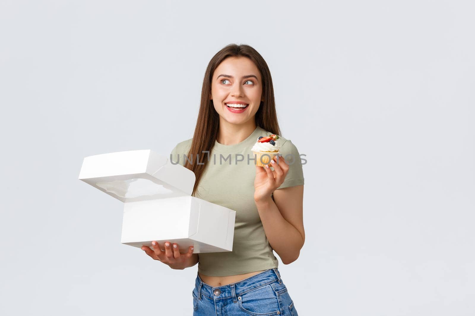 Delivery, lifestyle and food concept. Lovely good-looking woman enjoying eating dessert from cafe or pastry shop. Girl ordered cupcakes smiling pleased as like sweet food, white background.