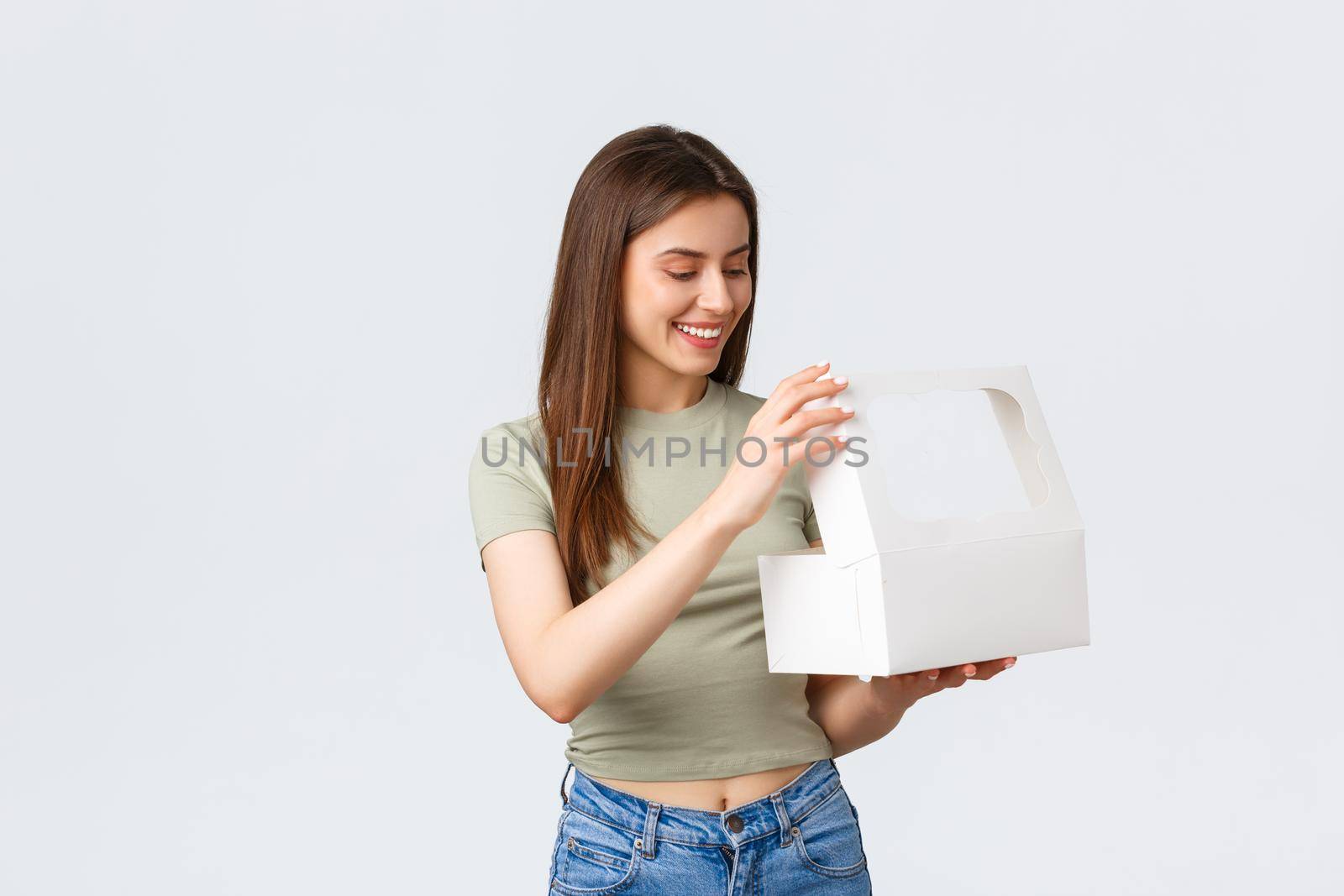 Delivery, lifestyle and food concept. Happy pleased female customer receive order from favourite cafe or restaurant, open white box with desserts, cake or muffins, white background.