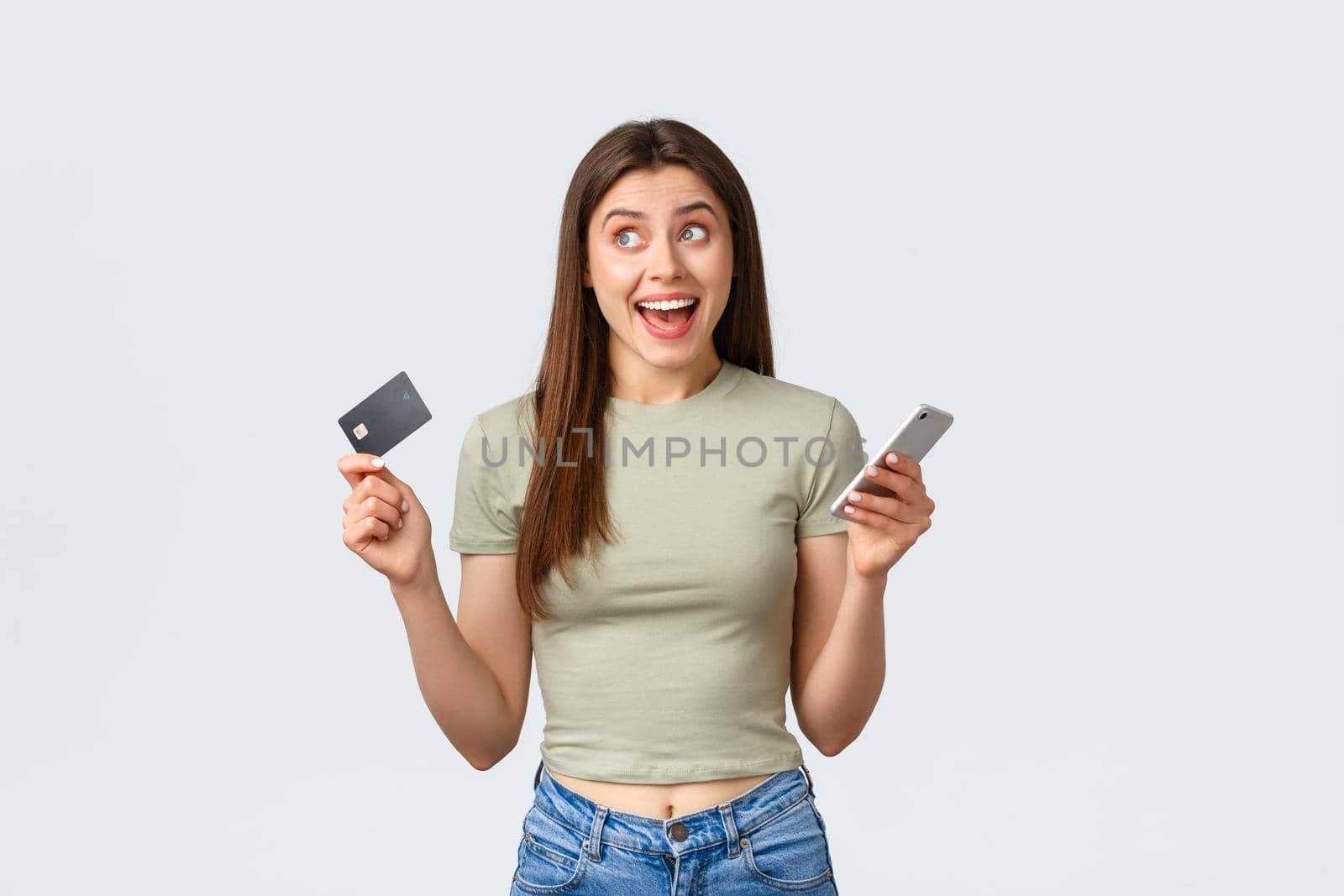 Online shopping, home lifestyle and people concept. Happy cheerful, smiling brunette woman dreaming of new dress, looking up with rejoice as holding credit card with mobile phone.