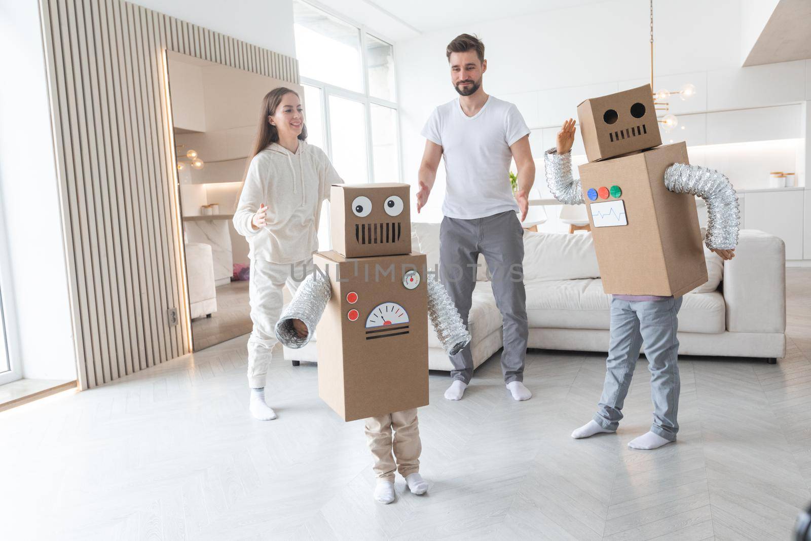 Happy family of parents and two children playing dancing like robots at home, children wearing handmade moving box costume of cardboard