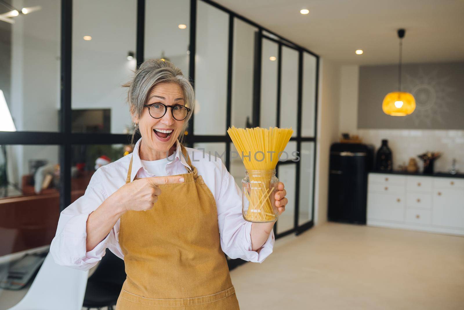 Happy senior woman showing pasta on camera. Portrait of smiling aged lady holding spaghetti standing in modern kitchen