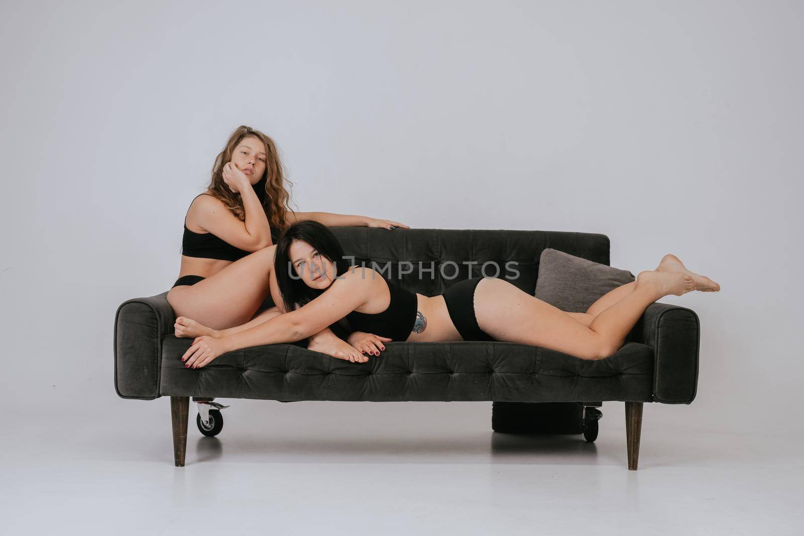 Two girlfriends wearing comfortable underwear on a big sofa, look at camera having smile and natural unique beauty