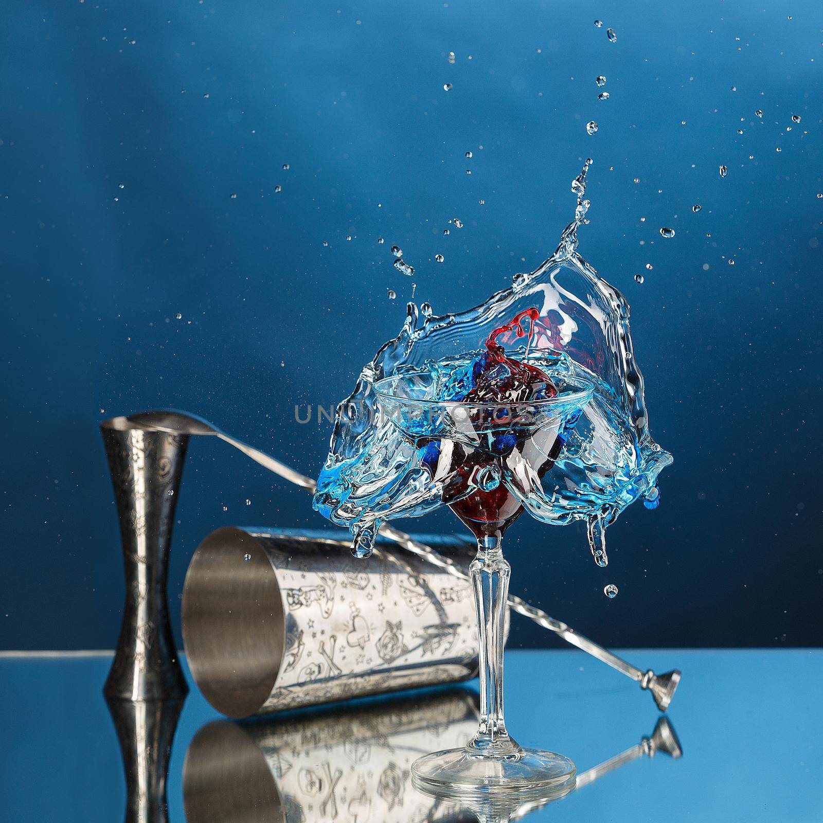 Splash from pouring martini into the glass. Cocktail, bar spoon and mixing glass on a blue background. Square image