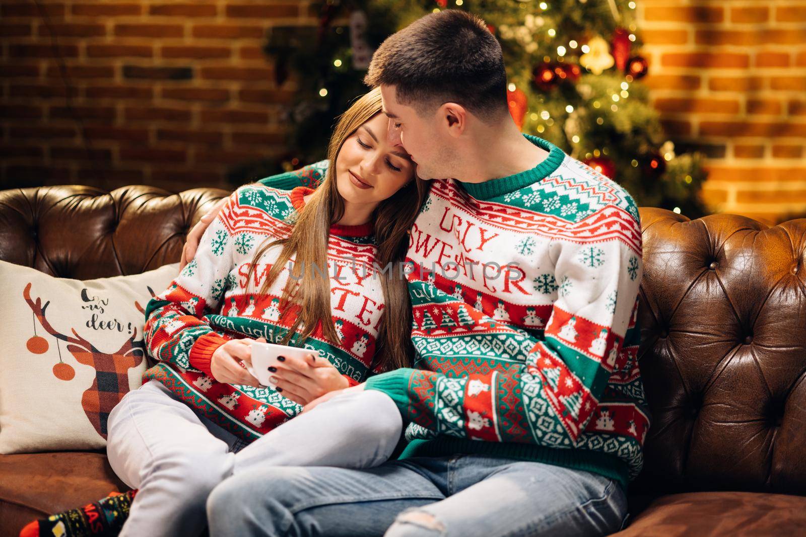 Happy couple enjoying Christmas together drinking hot tea. Couple Celebration New Year Near Christmas Tree. Christmas eve. Cute couple in love drinking hot tea and enjoying winter holidays at home