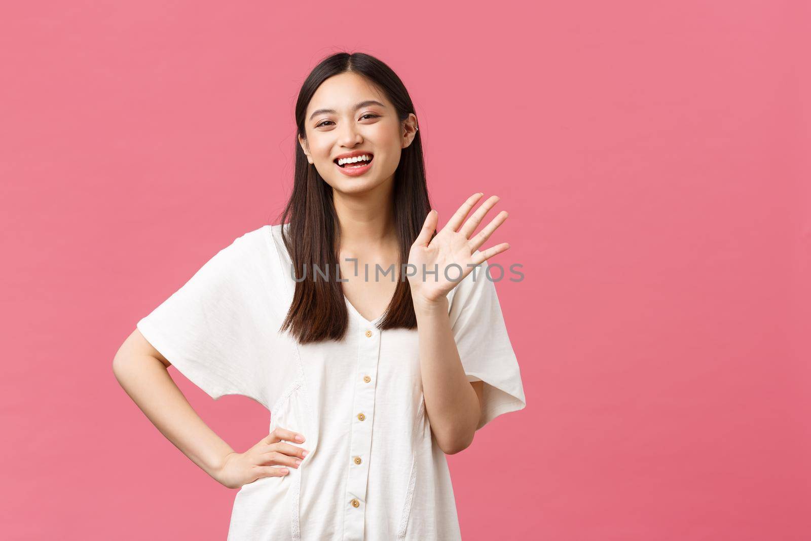 Beauty, people emotions and summer leisure concept. Friendly and outgoing pretty asian girl in white dress saying hi, waving hand and cheerful smiling, greeting someone hello gesture.