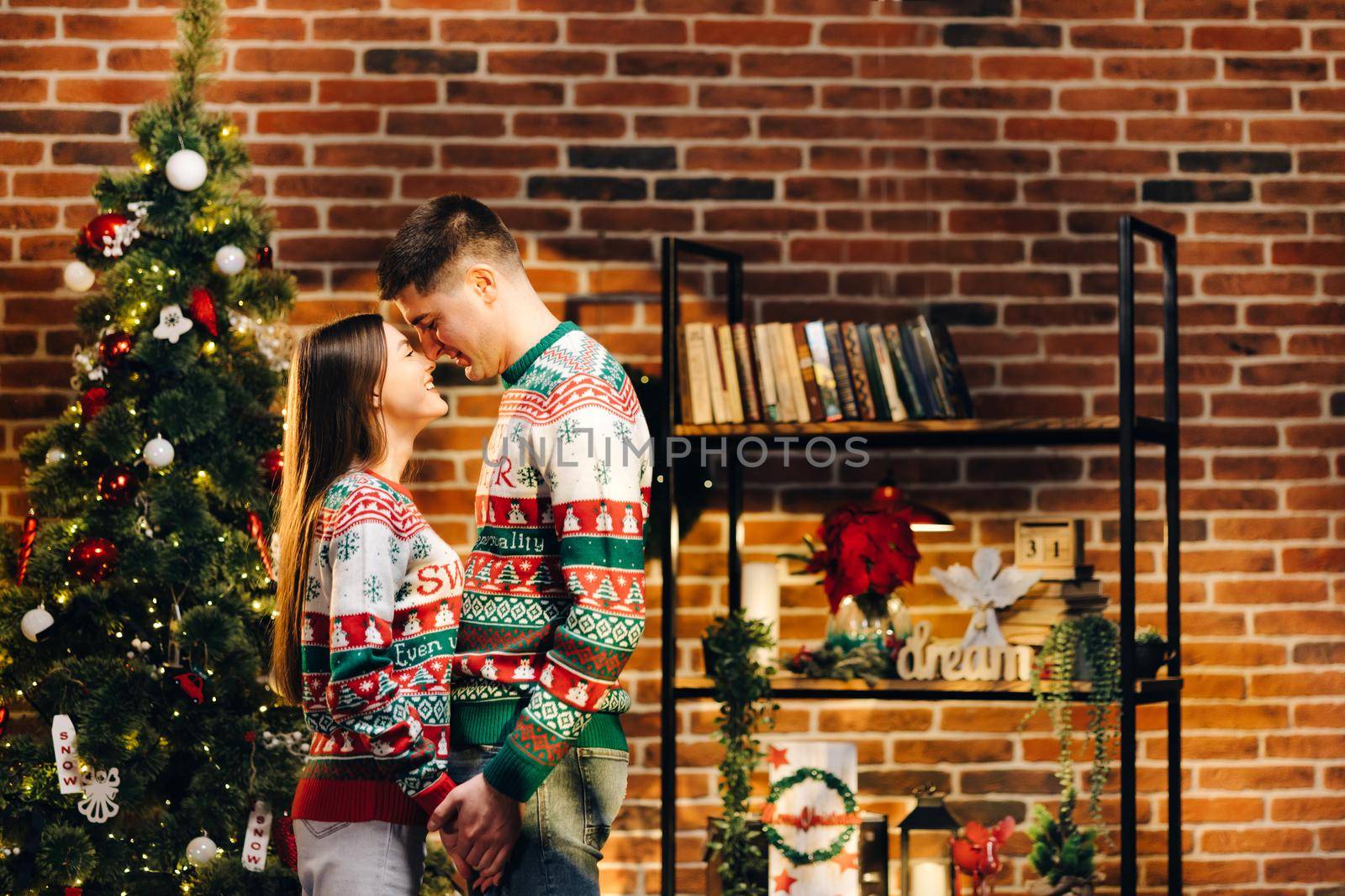Passionate romantic caucasian couple in sweaters are spending time together before New Year at home. Tender couple hugging on the background of shiny garland by uflypro