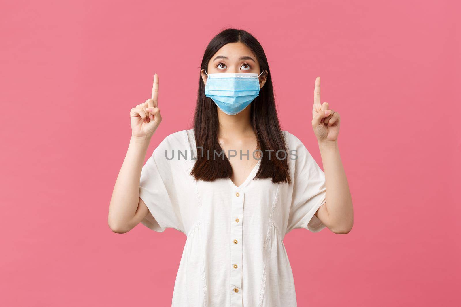 Covid-19, social distancing, virus and lifestyle concept. Curious pretty asian girl in medical mask and summer dress, looking and pointing fingers up at promo, advertisement, pink background.