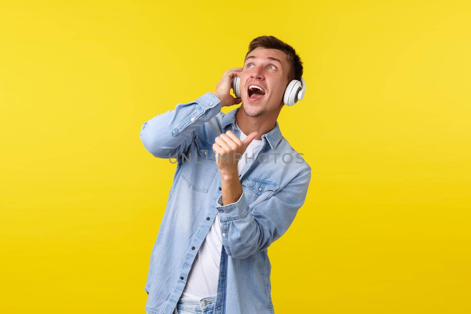 Lifestyle, summer holidays, technology concept. Handsome carefree male dj, guy enjoying awesome sound of music, showing thumbs-up at upper left corner, wearing wireless headphones, yellow background by Benzoix