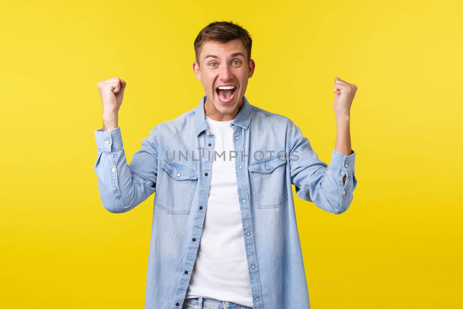 Lifestyle, people emotions and summer leisure concept. Excited handsome happy man rejoicing, raising hands up and chanting, rooting for team, being champion, triumphing over yellow background by Benzoix