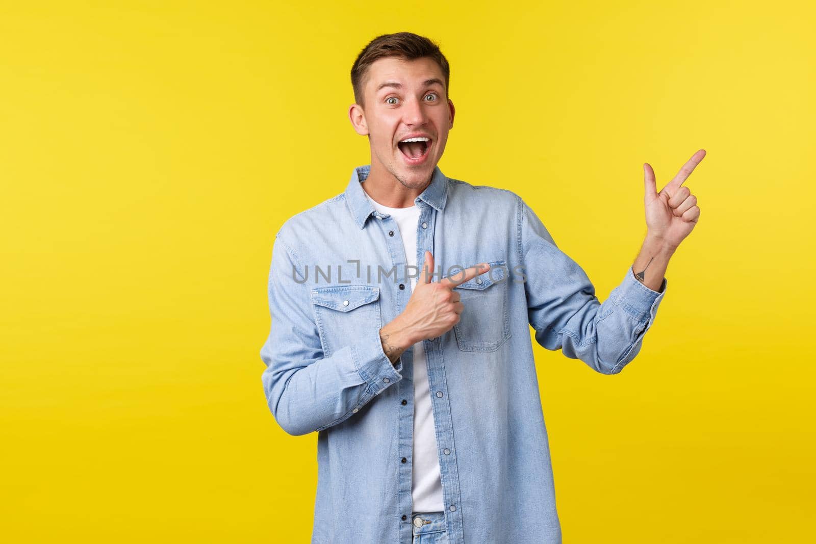 Good-looking happy man in casual clothes pointing fingers upper right corner and smiling excited, checking out awesome promo offer, demonstrate banner with advertisement, yellow background by Benzoix