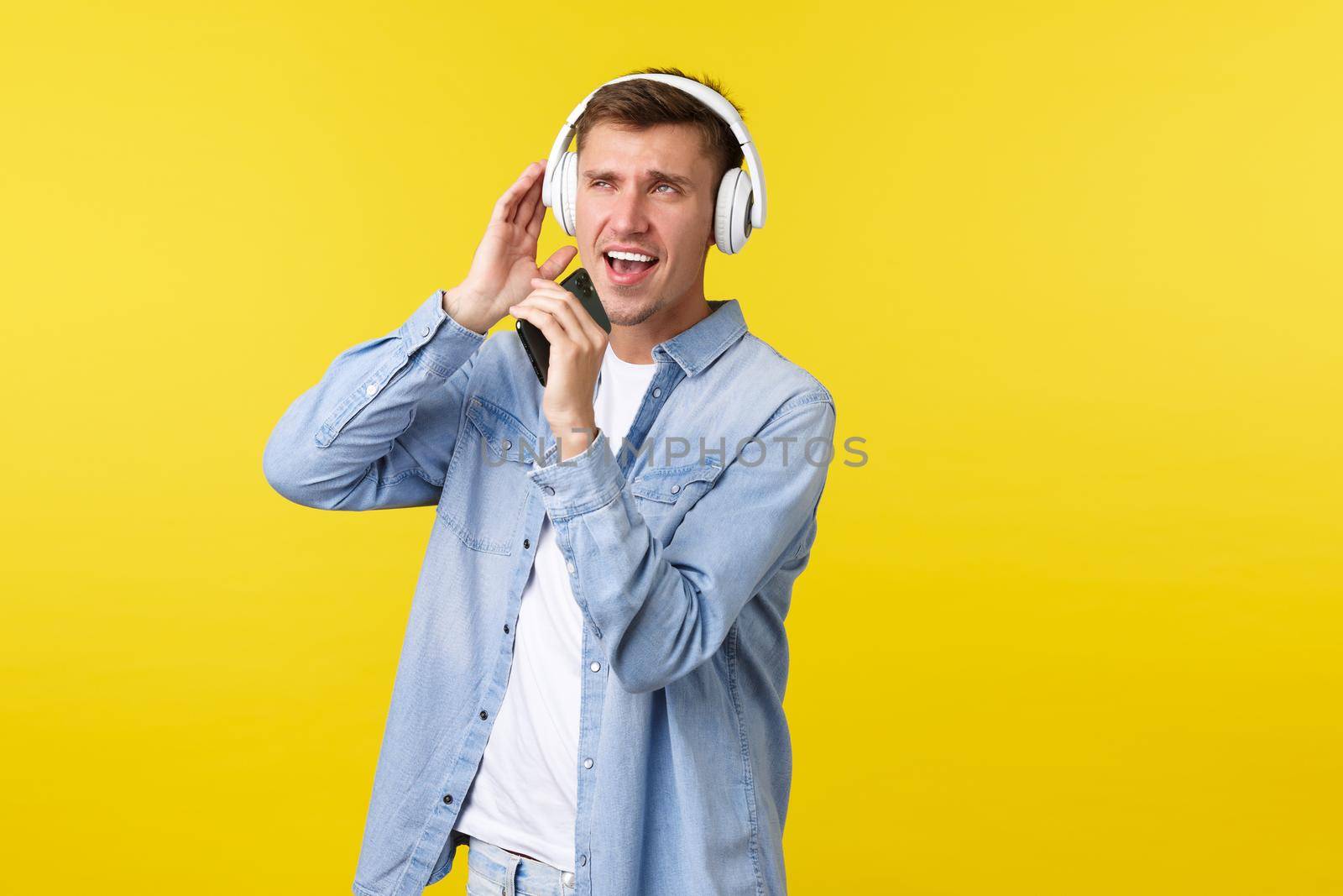 Lifestyle, summer holidays, technology concept. Happy good-looking blond guy in casual outfit, playing karaoke app, singing song into mobile phone and wearing headphones, yellow background.