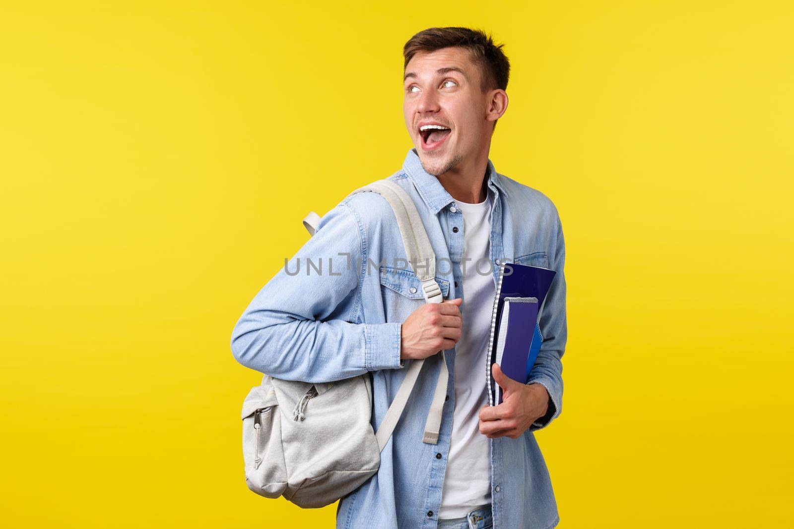 Education, courses and university concept. Excited happy smiling handsome student turn back at upper left corner while heading to classes with backpack and notebooks, yellow background.