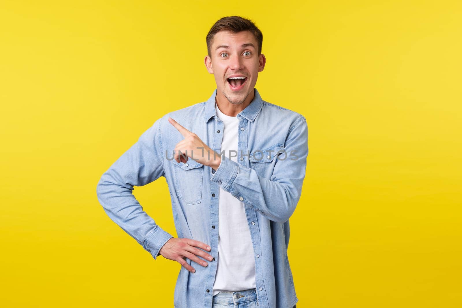 Enthusiastic happy handsome blond man in stylish outfit, pointing finger upper left corner and smiling broadly, found out amazing great news, showing special offer banner, yellow background by Benzoix