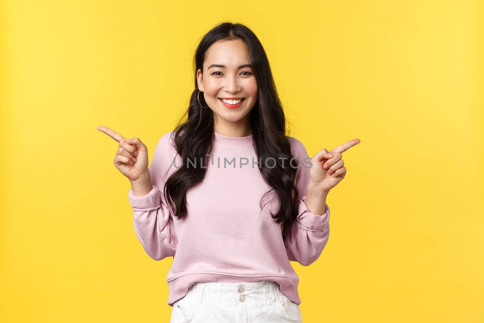 People emotions, lifestyle and fashion concept. Cheerful asian woman smiling and showing both products, pointing fingers sideways at two offers or banners over yellow background by Benzoix