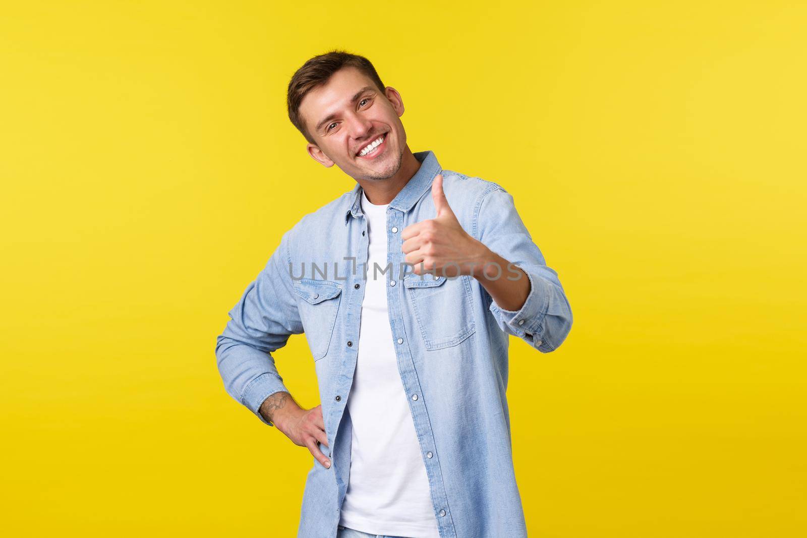 Portrait of satisfied proud young handsome man congratulate person for accomplishing goal, show thumbs-up delighted, smiling pleased. Man recommending awesome offer, stand yellow background by Benzoix