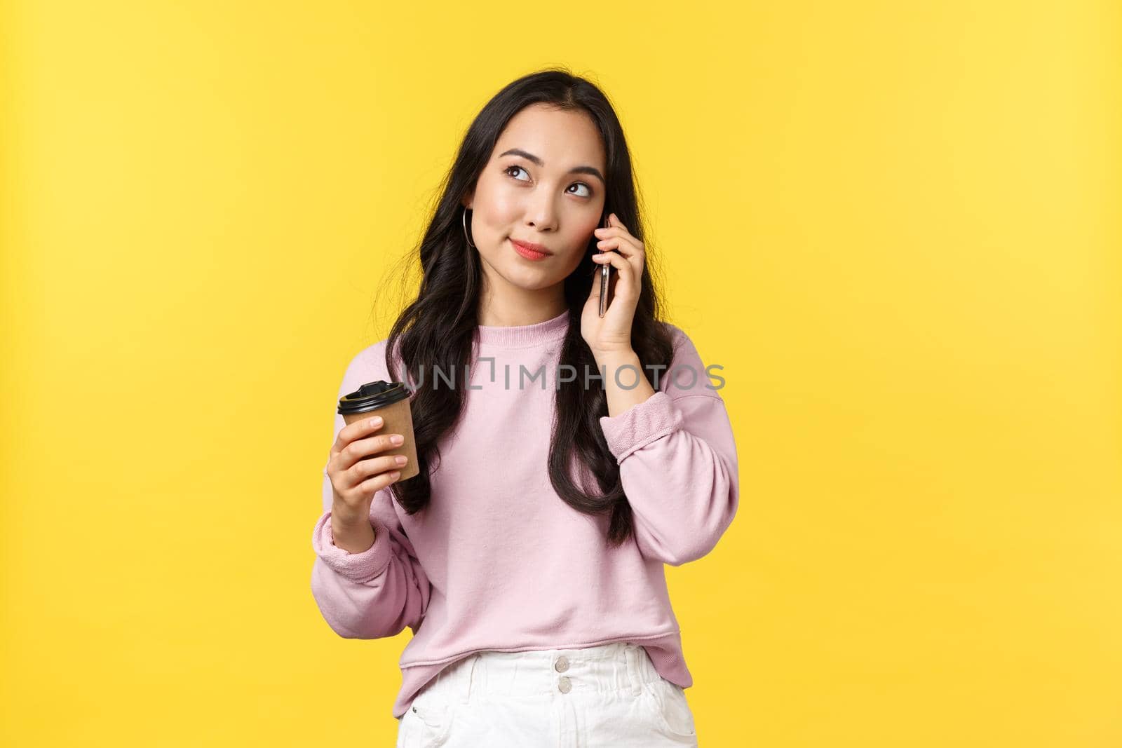 People emotions, lifestyle leisure and beauty concept. Cute stylish asian woman looking up thoughtful as talking on mobile phone and drinking coffee from takeaway cup, yellow background by Benzoix