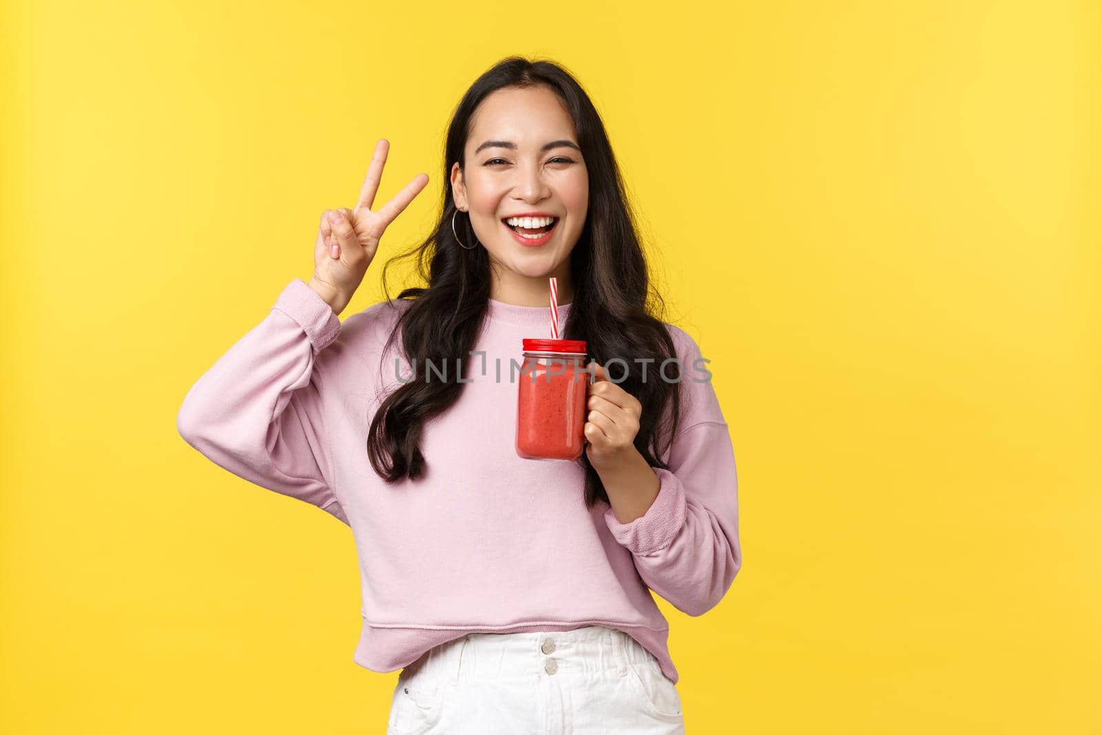 People emotions, lifestyle leisure and beauty concept. Happy upbeat asian 20s girl showing peace sign and smiling delighted as enjoying summer drink, holding smoothie, yellow background.