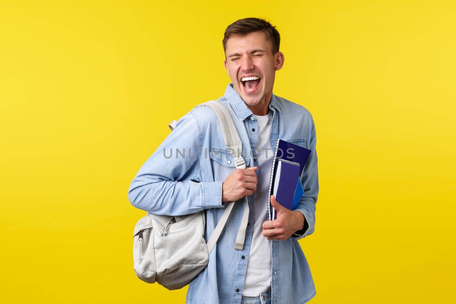 Education, courses and university concept. Joyful happy handsome male student enjoying life in campus, holding backpack and study material, laughing and smiling excited, yellow background.