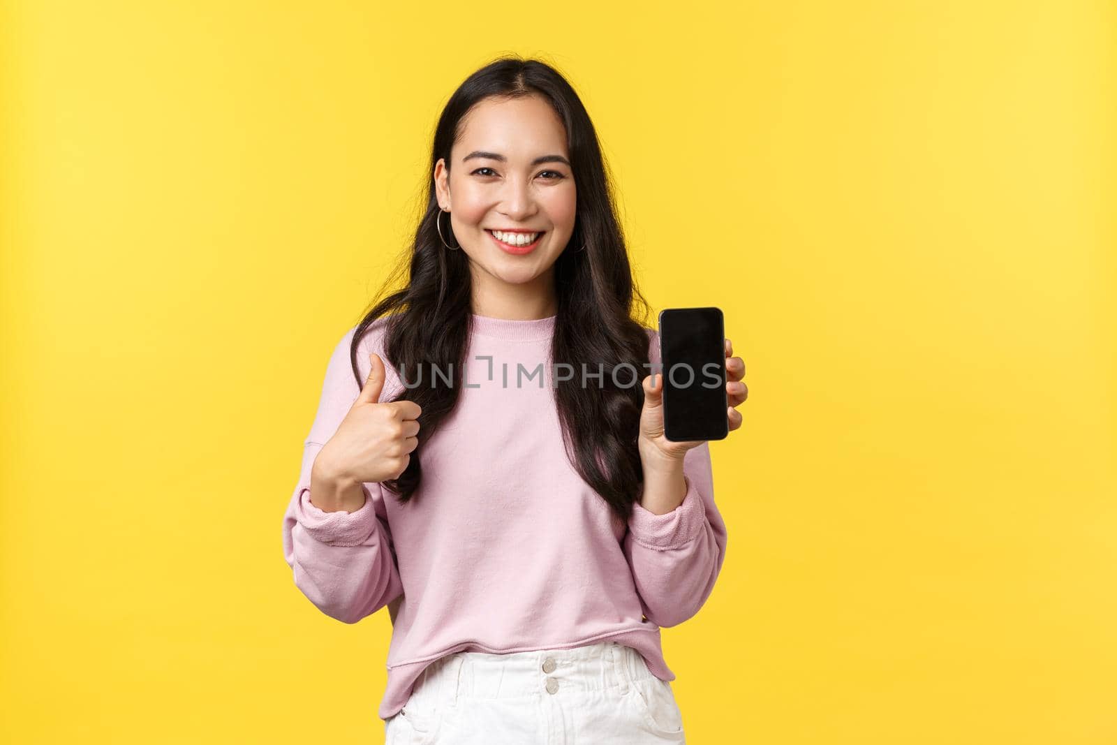 People emotions, lifestyle leisure and beauty concept. Smiling asian woman 20s, showing smartphone display, recommend application or mobile game, showing thumbs-up in approval, yellow background.