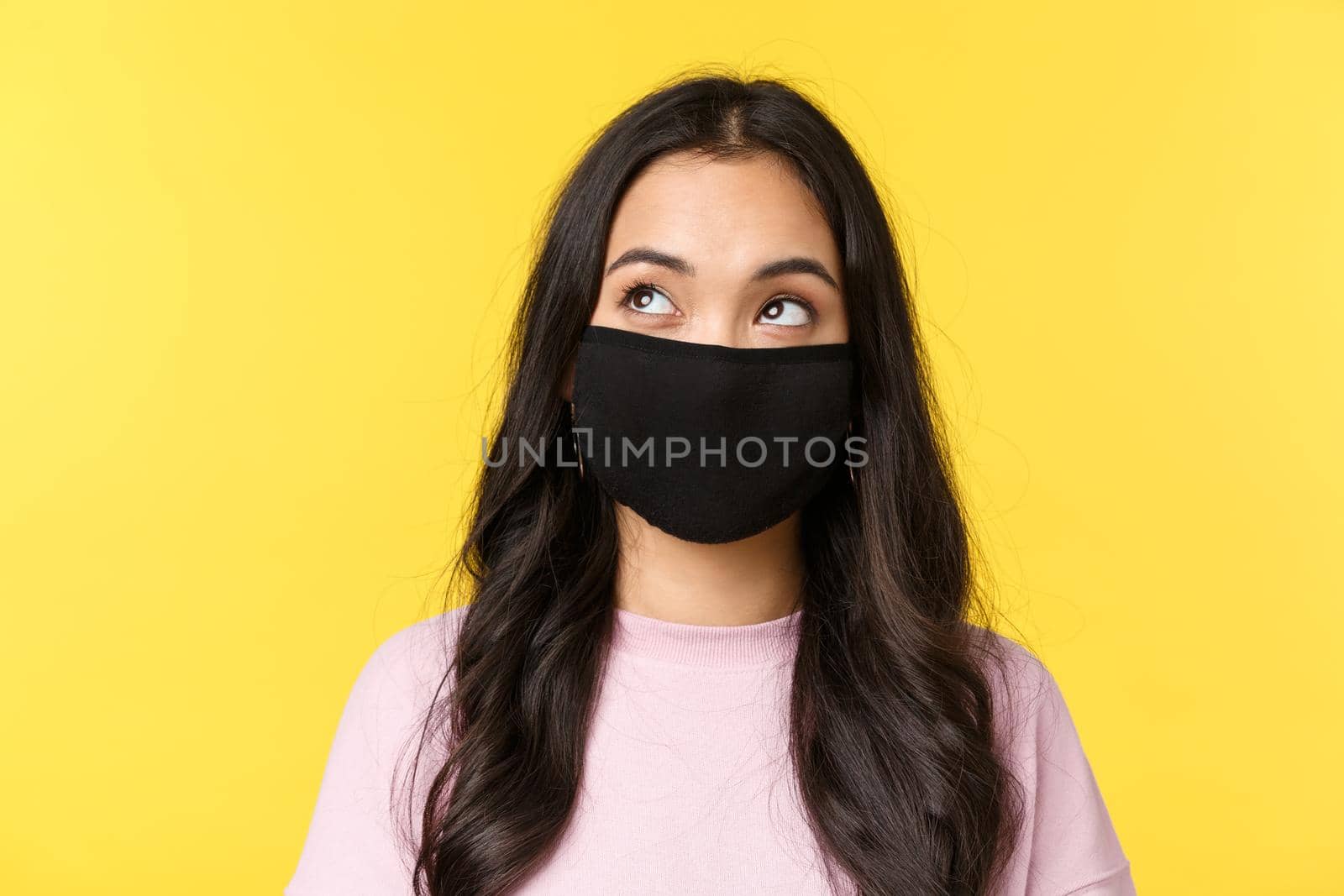 Covid-19, social-distancing lifestyle, prevent virus spread concept. Dreamy and thoughtful asian girl in face mask, looking upper left corner thinking, standing yellow background.
