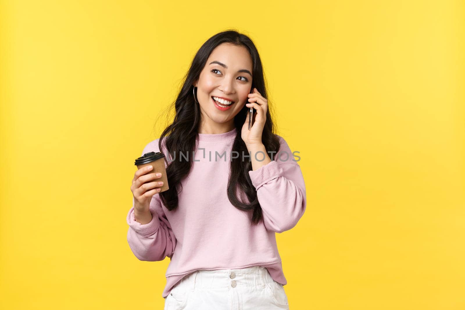 People emotions, lifestyle leisure and beauty concept. Modern cute asian girl talking on phone with friend and smiling, drinking coffee casually, hold takeaway cup, standing yellow background.
