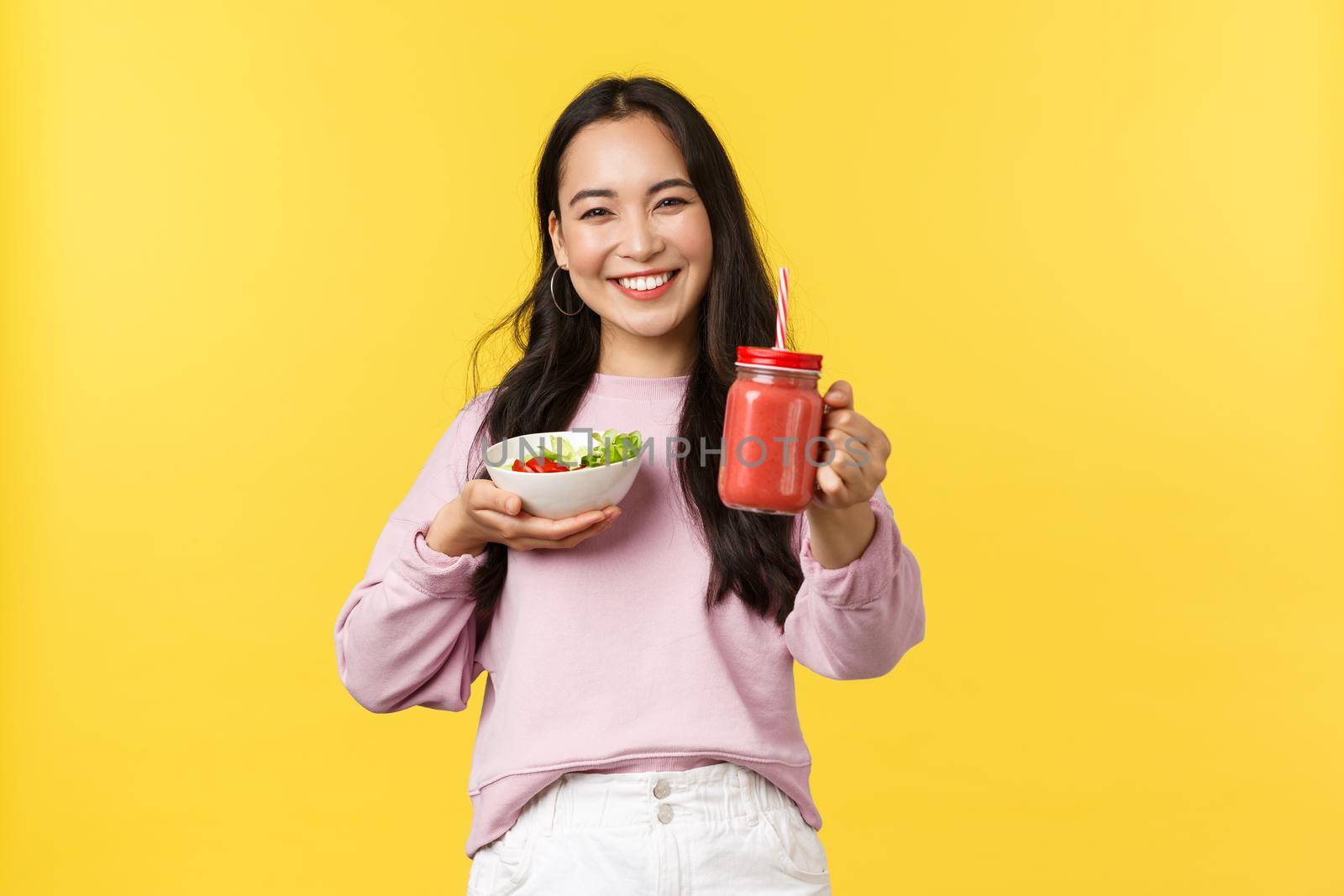 People emotions, healthy lifestyle and food concept. Friendly and cheerful smiling asian girl extend hand and give you smoothie, holding salad, sharing her lunch to eat together, yellow background.