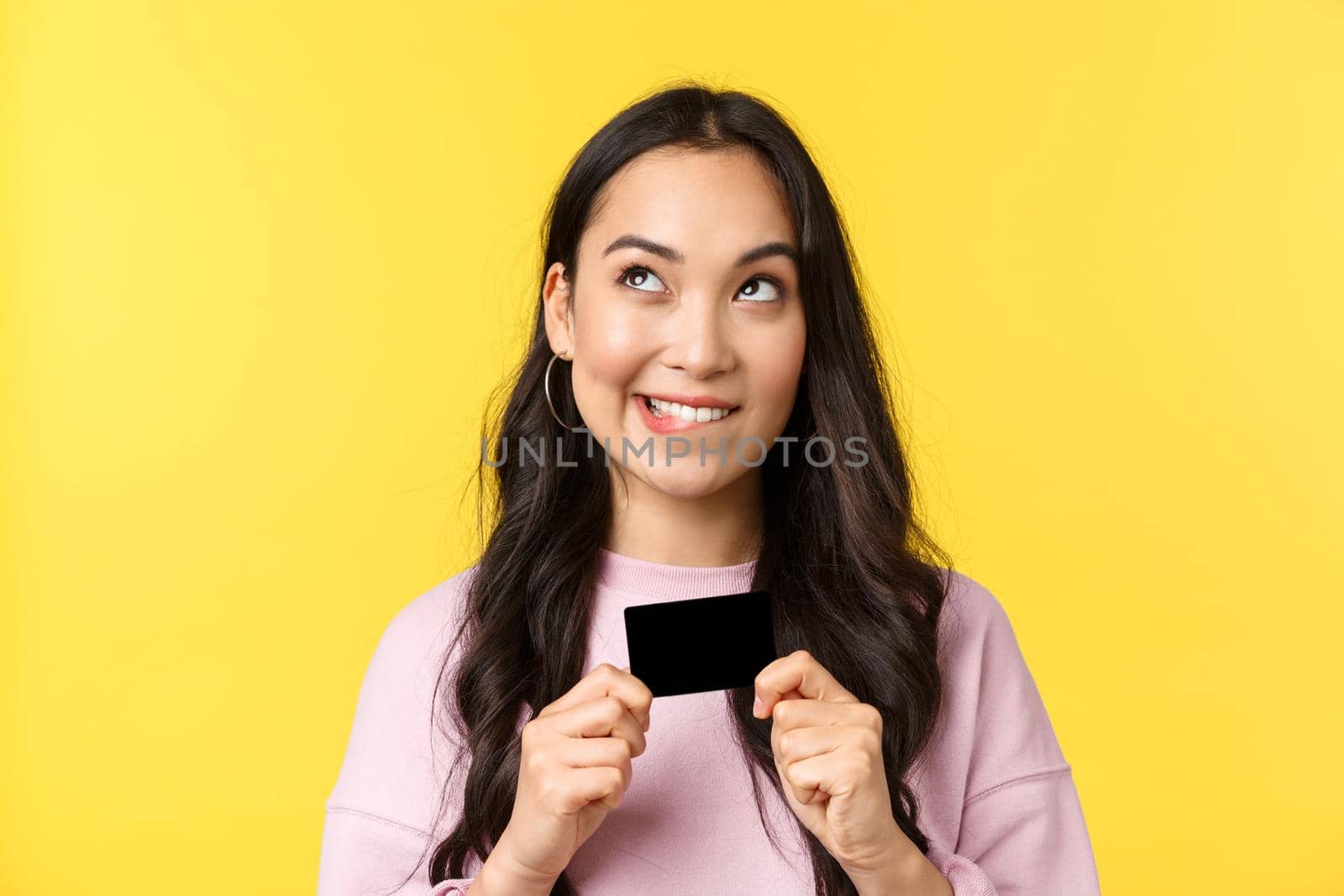 People emotions, lifestyle leisure and beauty concept. Dreamy asian girl thinking what buy next, looking upper left corner and smiling, holding credit card, going online shopping, yellow background.