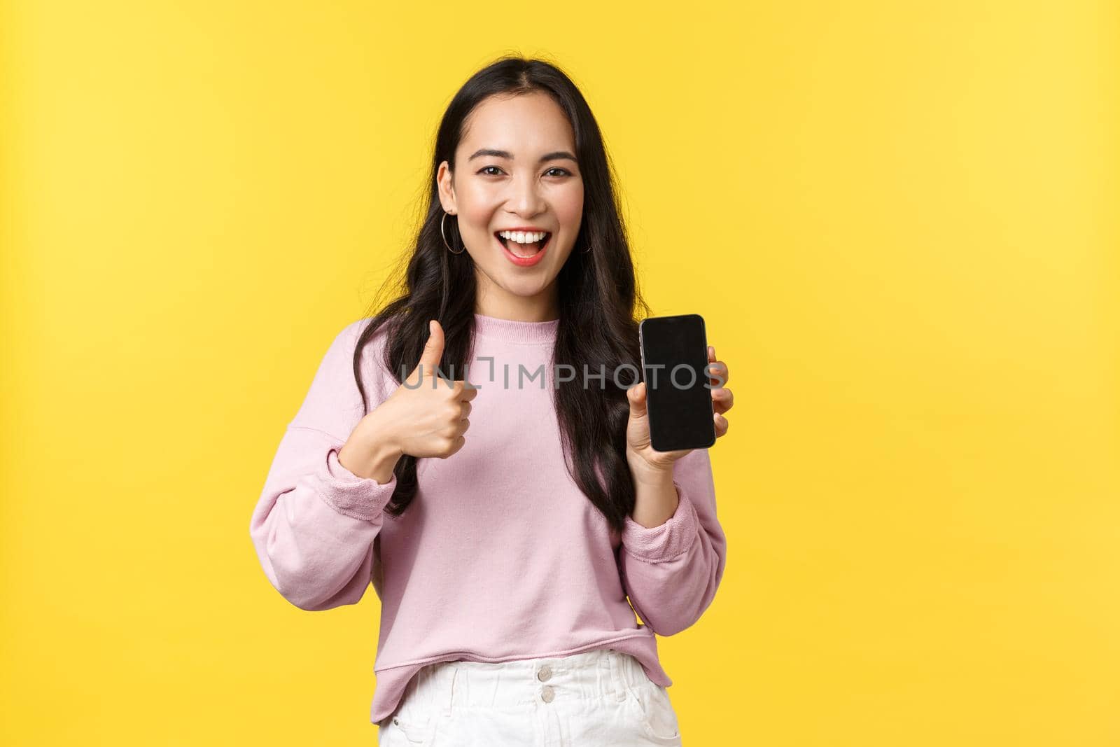 People emotions, lifestyle leisure and beauty concept. Smiling asian woman 20s, showing smartphone display, recommend application or mobile game, showing thumbs-up in approval, yellow background by Benzoix